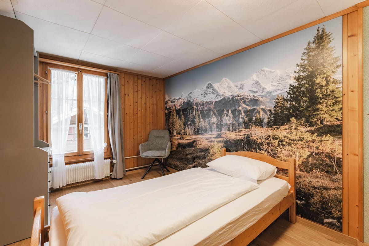Une chambre avec un lit en bois, une chaise et une grande fresque murale représentant un paysage de montagne.