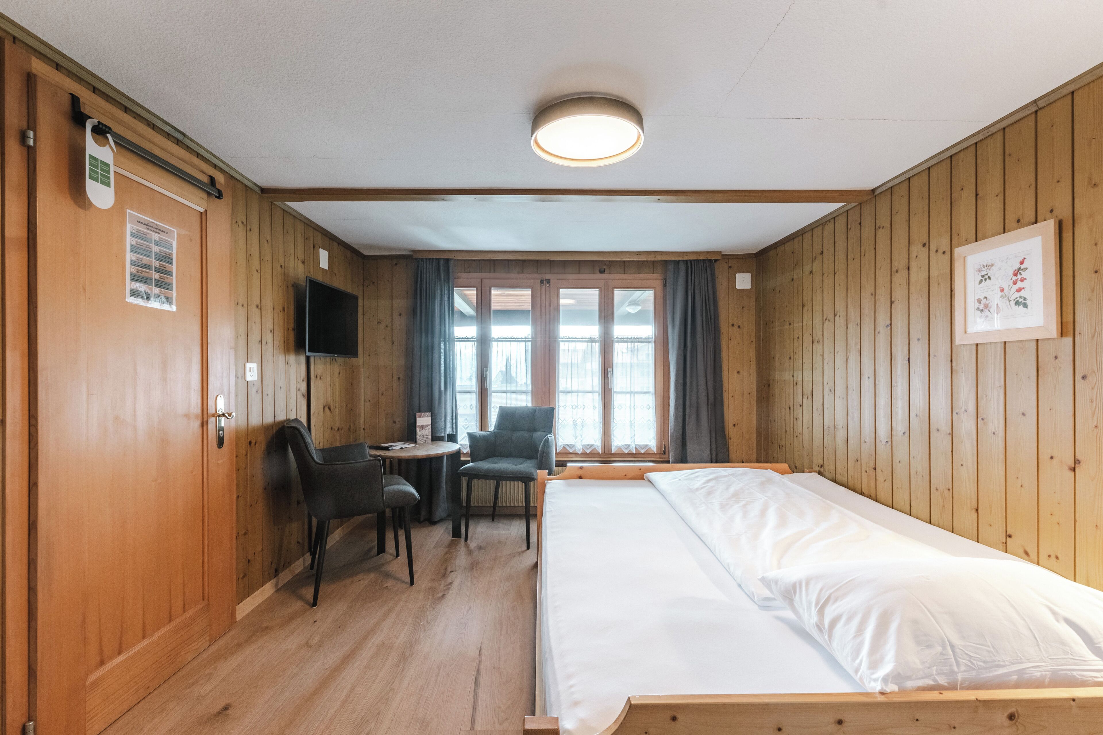 A room with wooden walls, a bed, a desk with a chair, a TV, and a window with curtains.