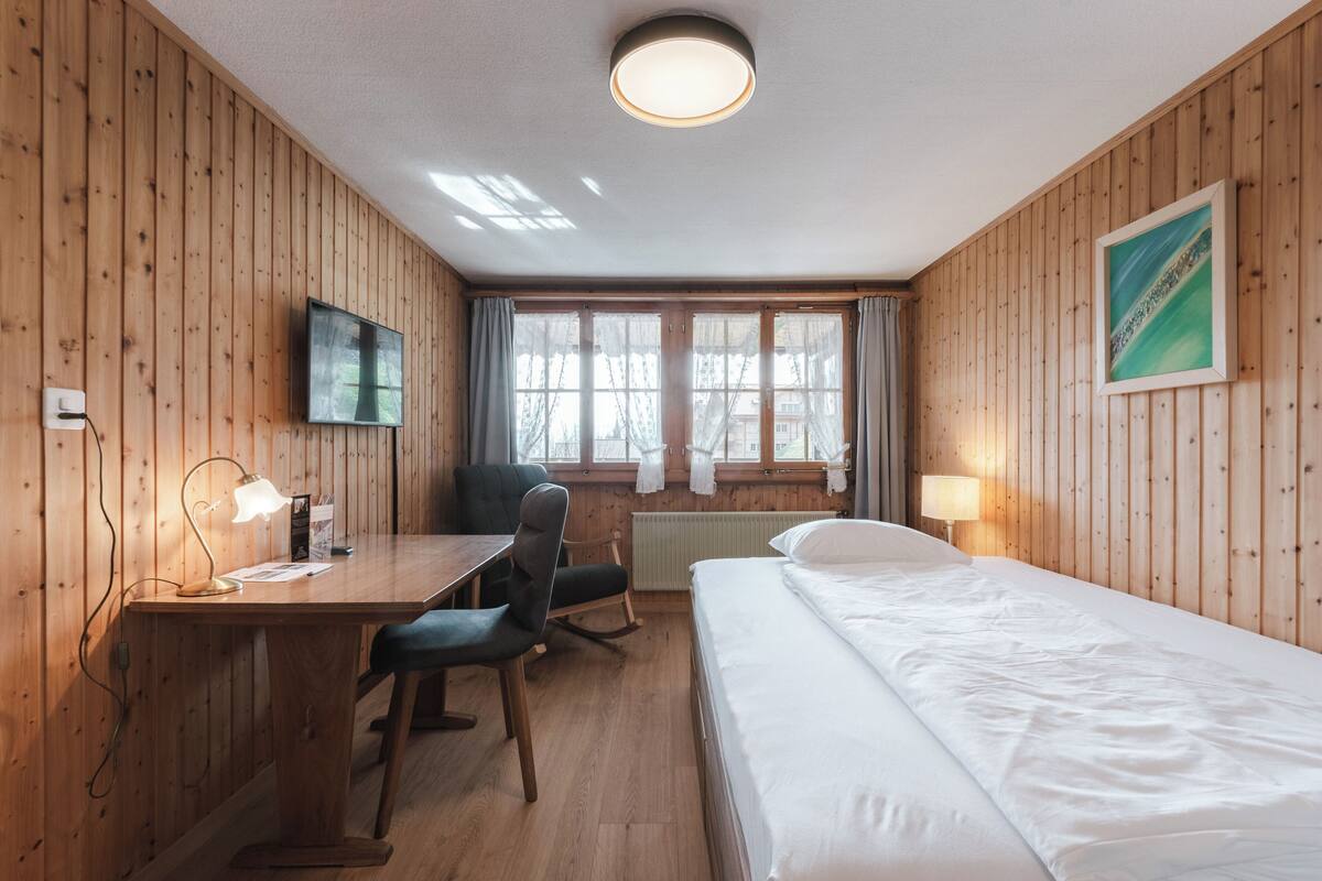 Une pièce aux murs lambrissés, comprenant un lit, un bureau, une chaise, un téléviseur et des fen êtres.