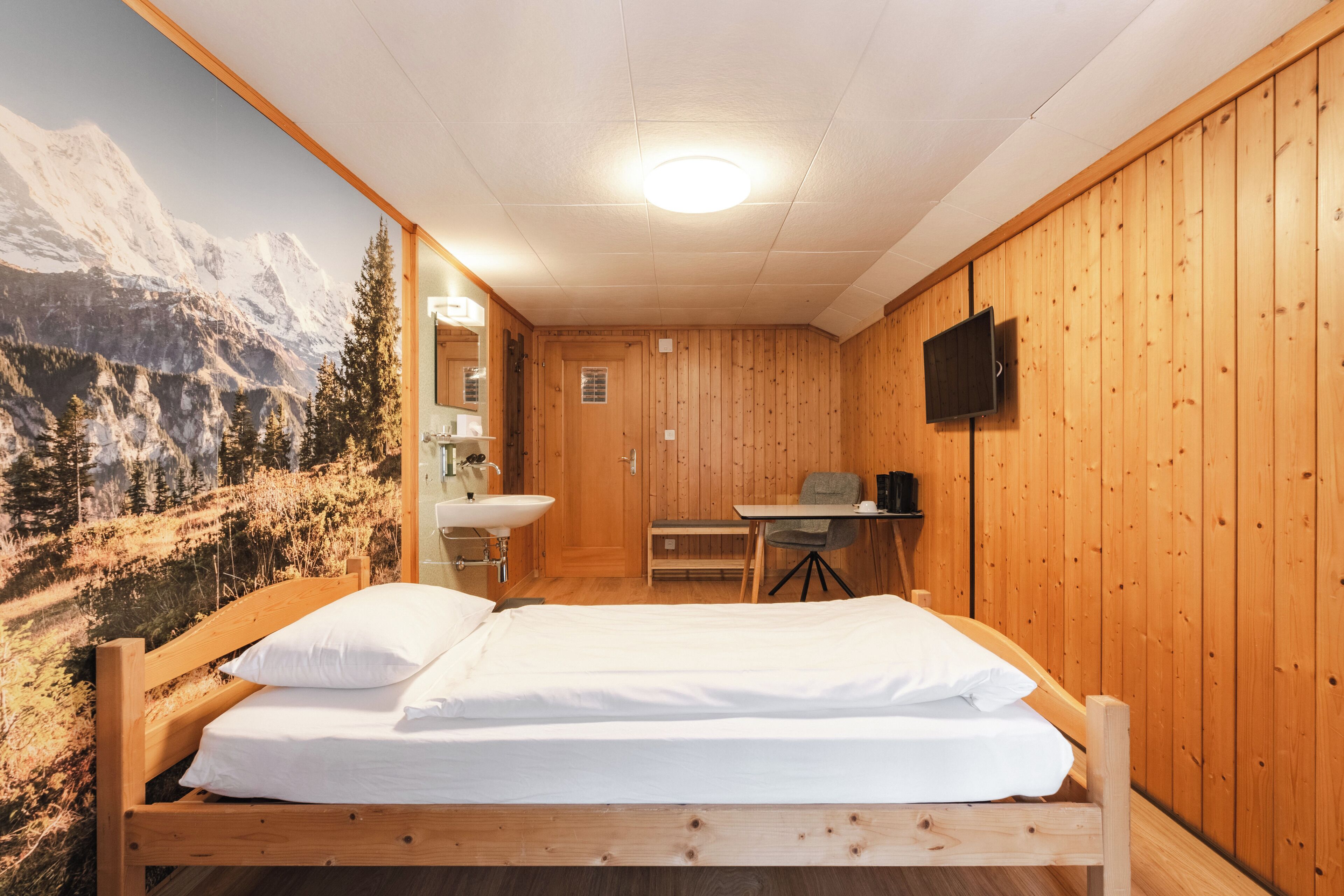 Una habitación de madera con cama, escritorio, silla, lavamanos y televisor.