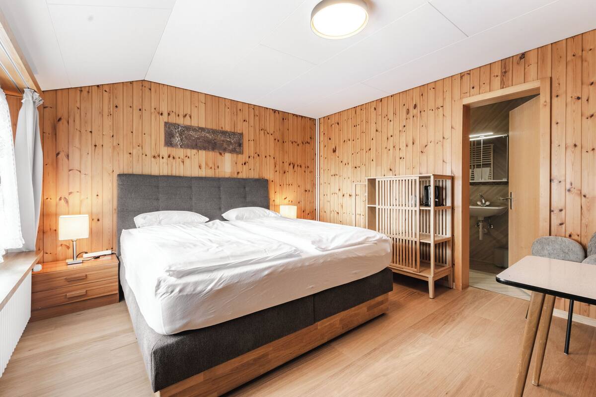 Une chambre aux murs lambrissés, avec un grand lit, des lampes de chevet, une armoire en bois et une petite table.
