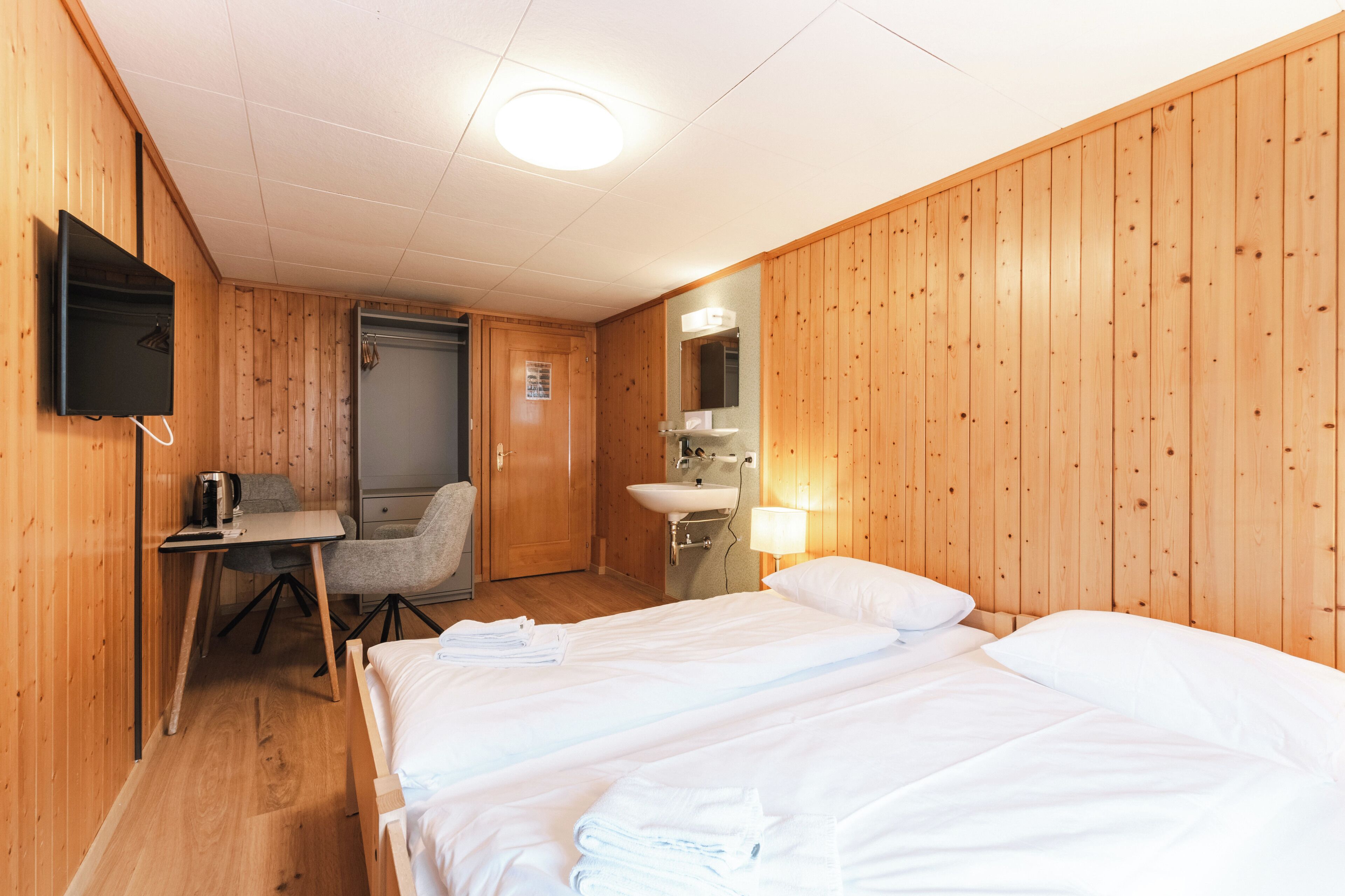 Una habitación con paredes de madera, una cama con ropa blanca, un escritorio con una silla, un lavamanos y un televisor montado en la pared.