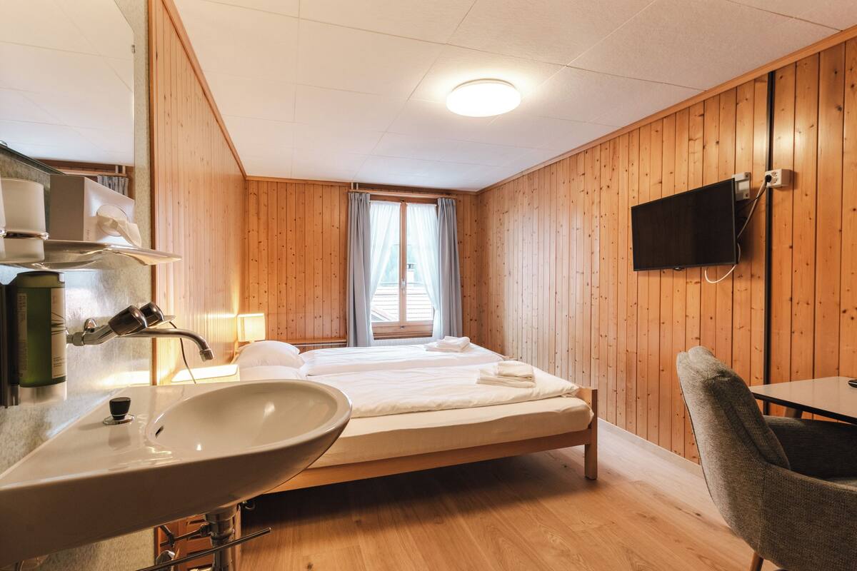 Une chambre d’hôtel avec des murs en bois, un grand lit, une table de chevet, un téléviseur fixé au mur et une salle de bain équipée d’un lavabo et d’un miroir.