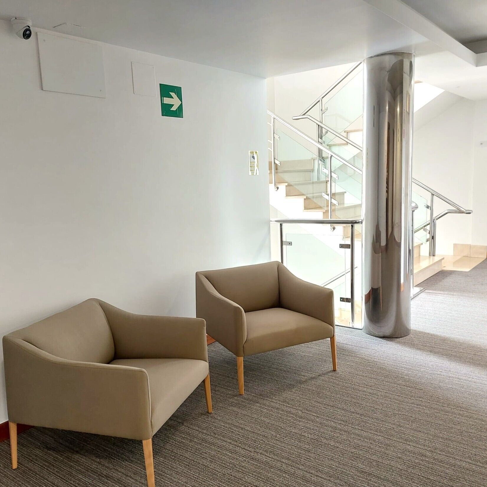 Un hall d’entrée moderne avec deux fauteuils beiges, un escalier en verre et un panneau de signalisation vert.