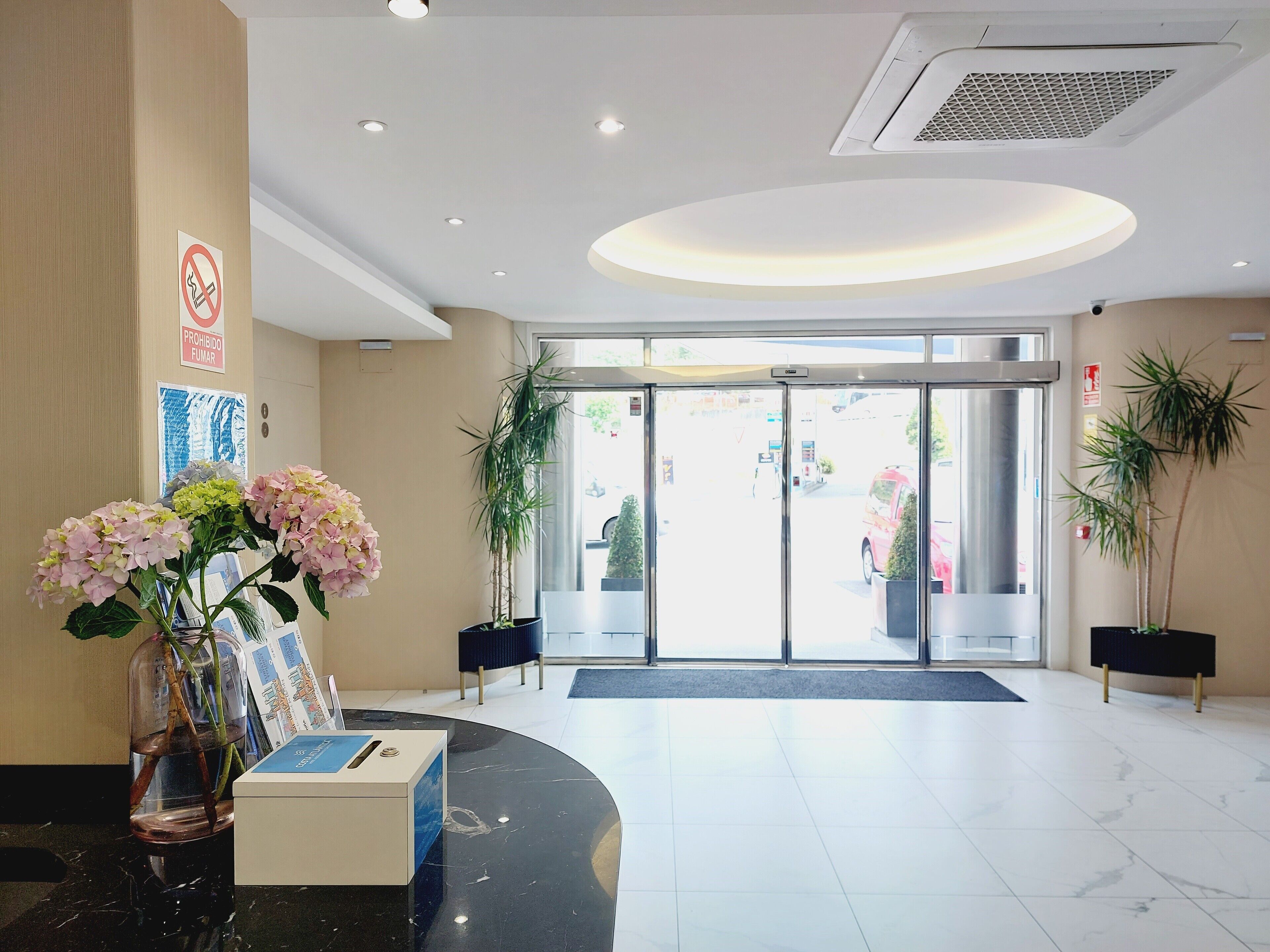 A hotel lobby with a reception desk, a no-smoking sign, a large glass door entrance, and potted plants.