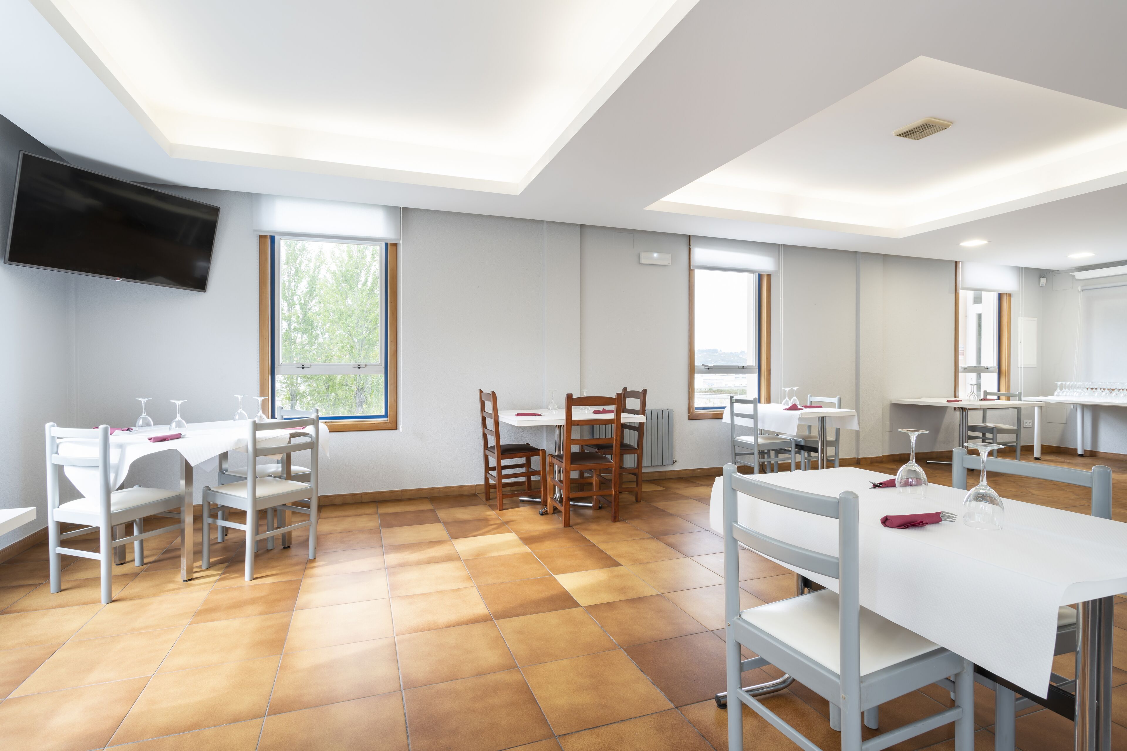 A spacious dining area with multiple tables and chairs, large windows, and a flat-screen TV mounted on the wall.