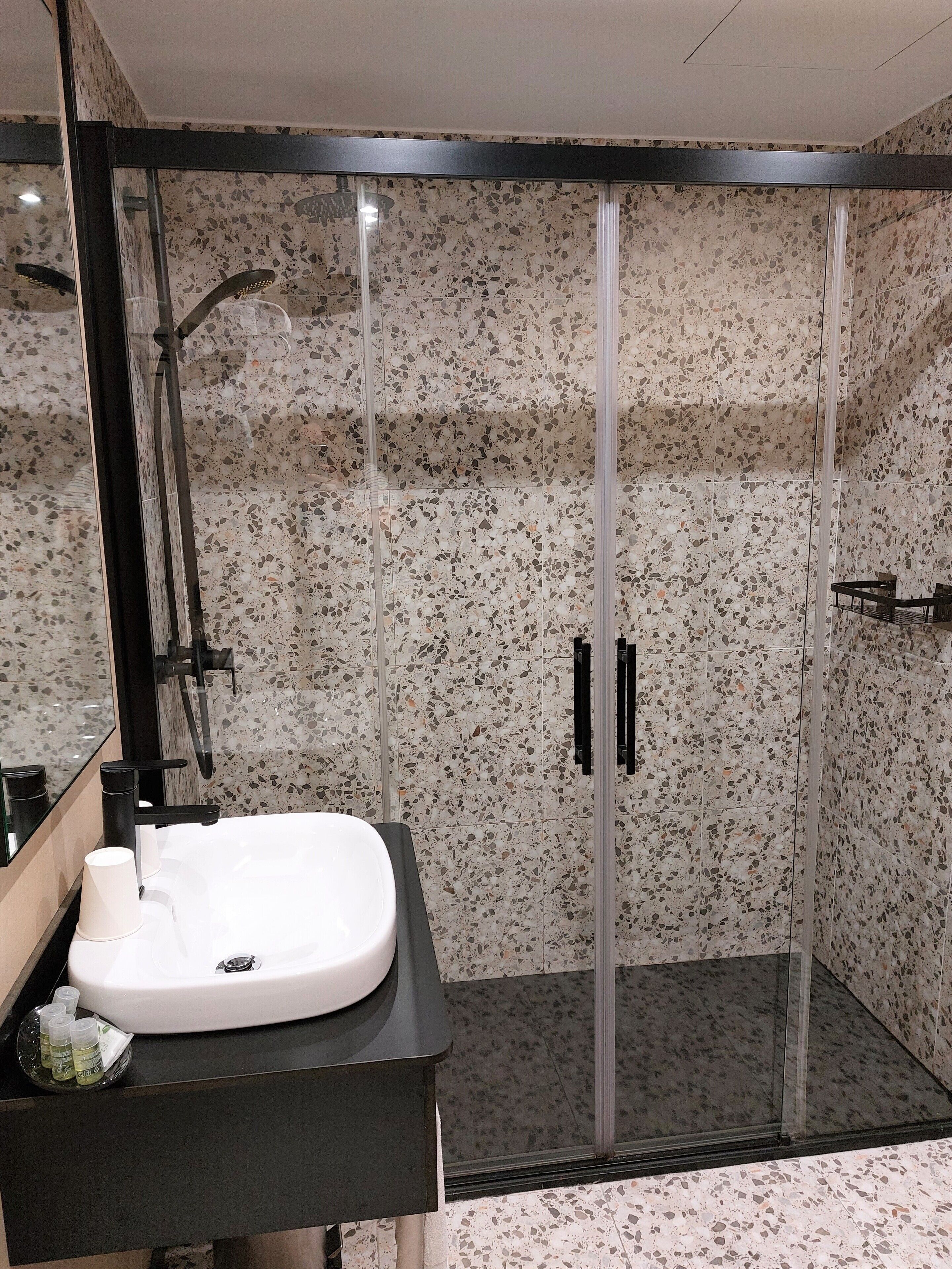 A modern bathroom with a glass-enclosed shower, a white sink with a black countertop, and a speckled tile backsplash.