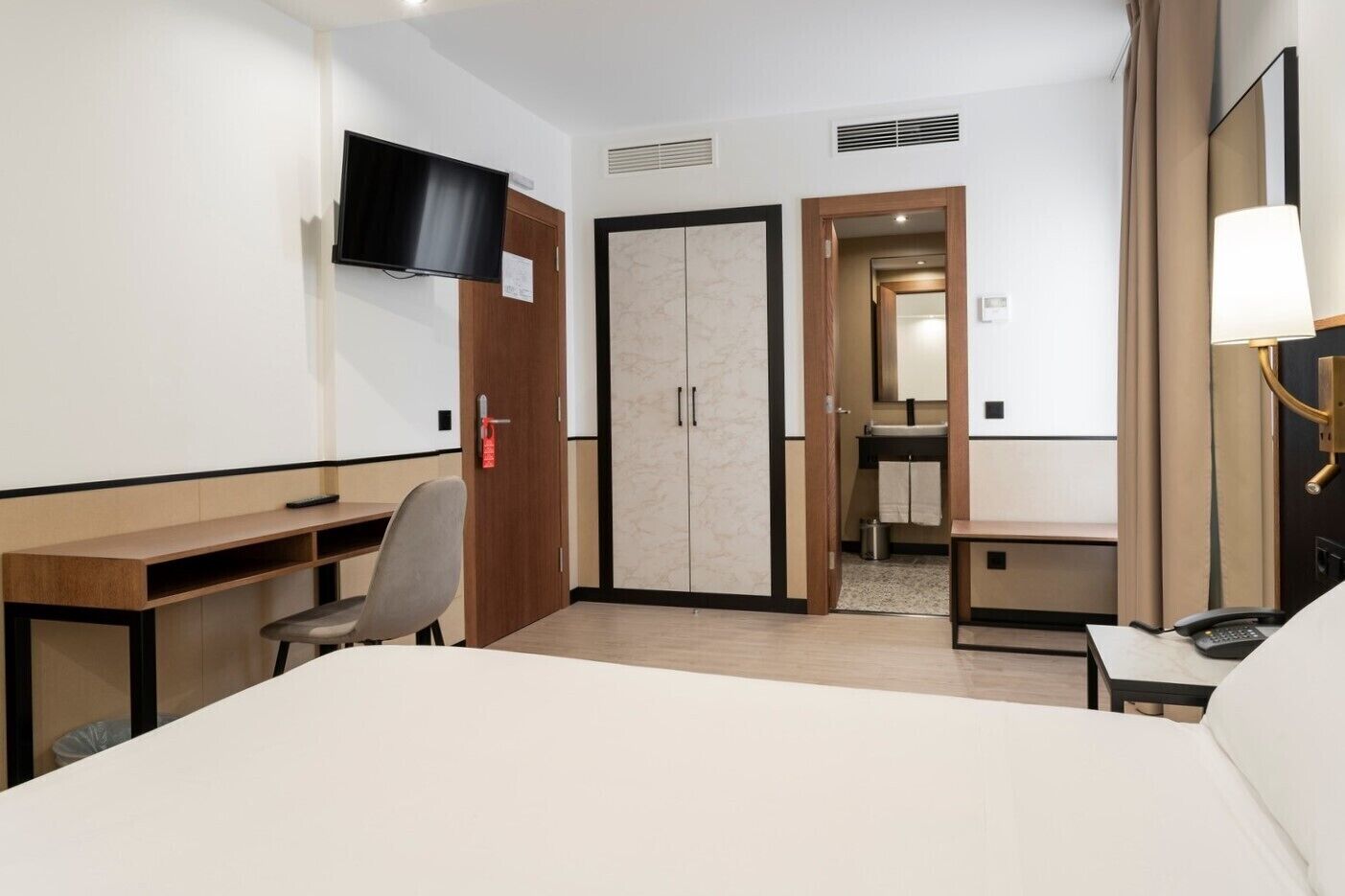 Habitación de hotel con cama, escritorio, silla, televisor, armario y baño visible a través de una puerta abierta.
