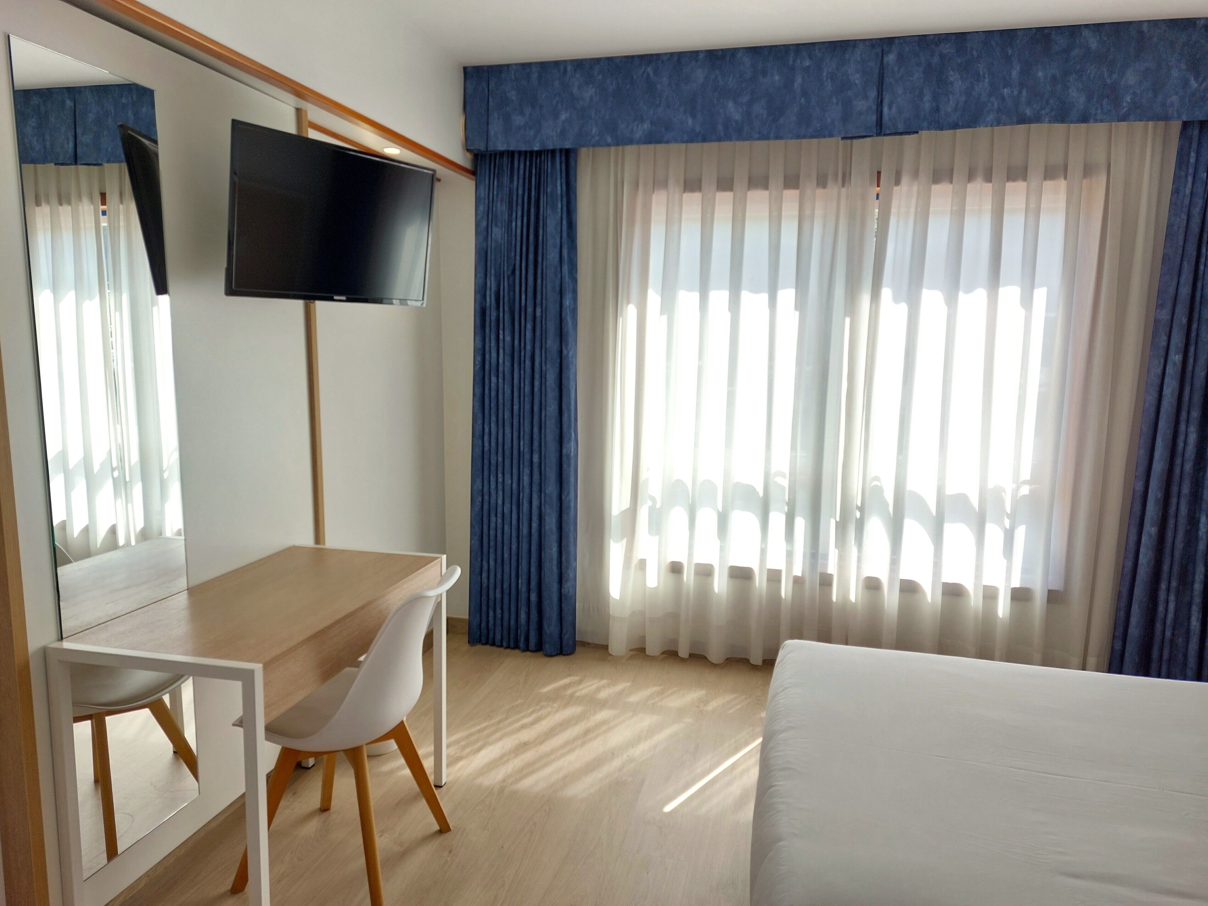 Ein Hotelzimmer mit Bett, Schreibtisch, Stuhl, Fernseher und einem Fenster mit Vorhängen.
