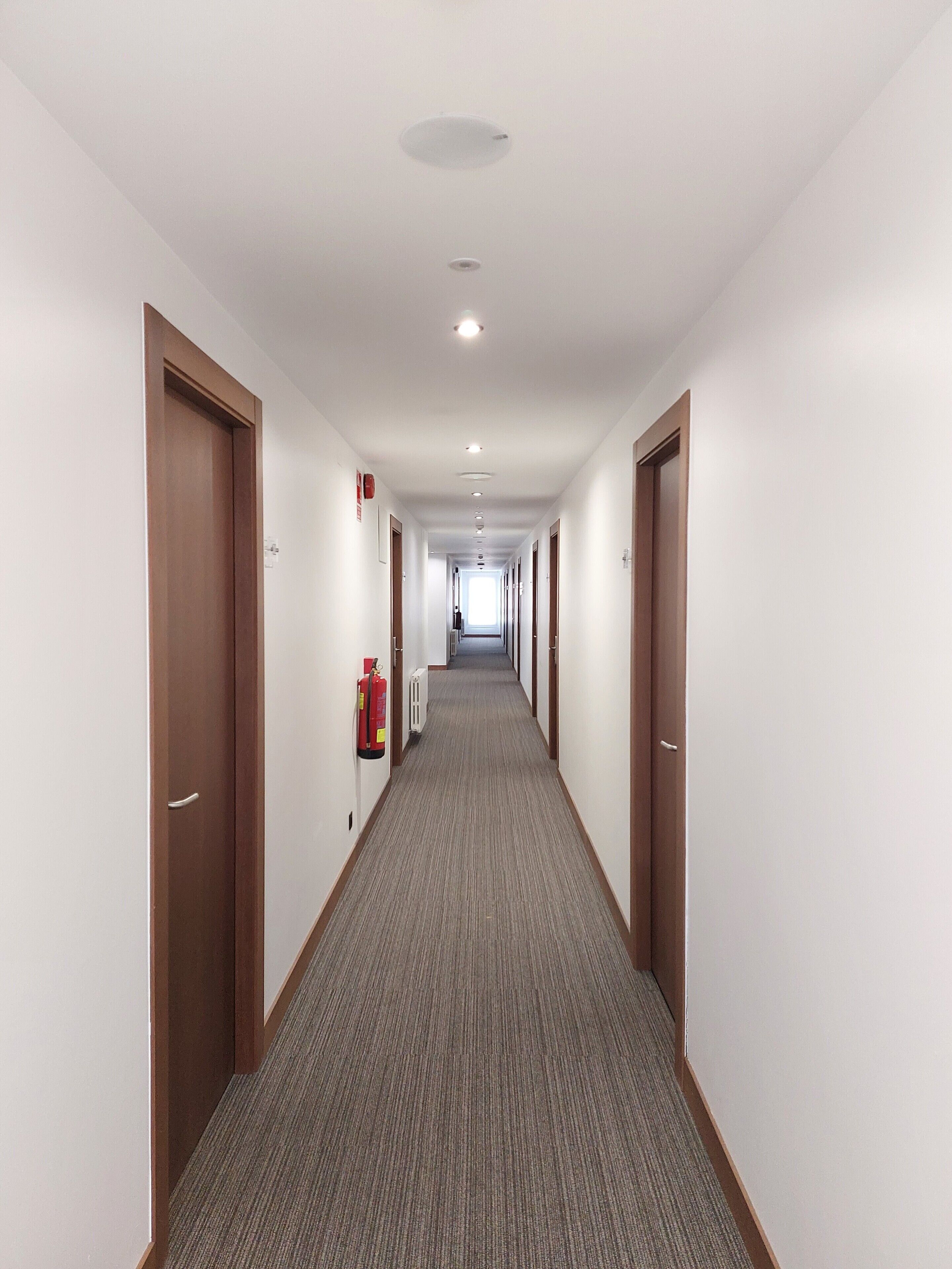 Un largo y estrecho pasillo de hotel con puertas de madera a ambos lados, suelo alfombrado y un extintor de incendios montado en la pared.