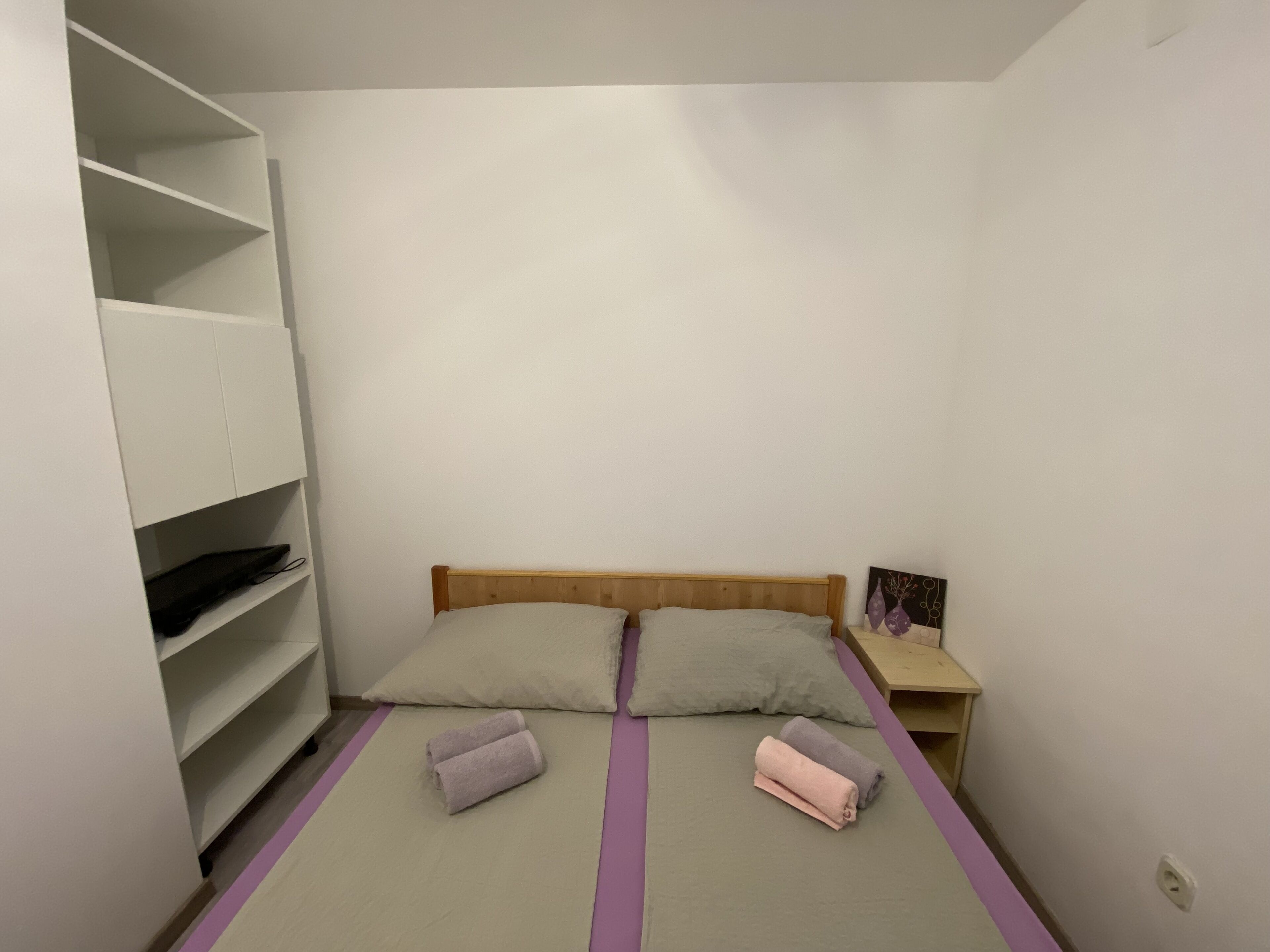A small, simple room with a bed, a nightstand, and a shelving unit.