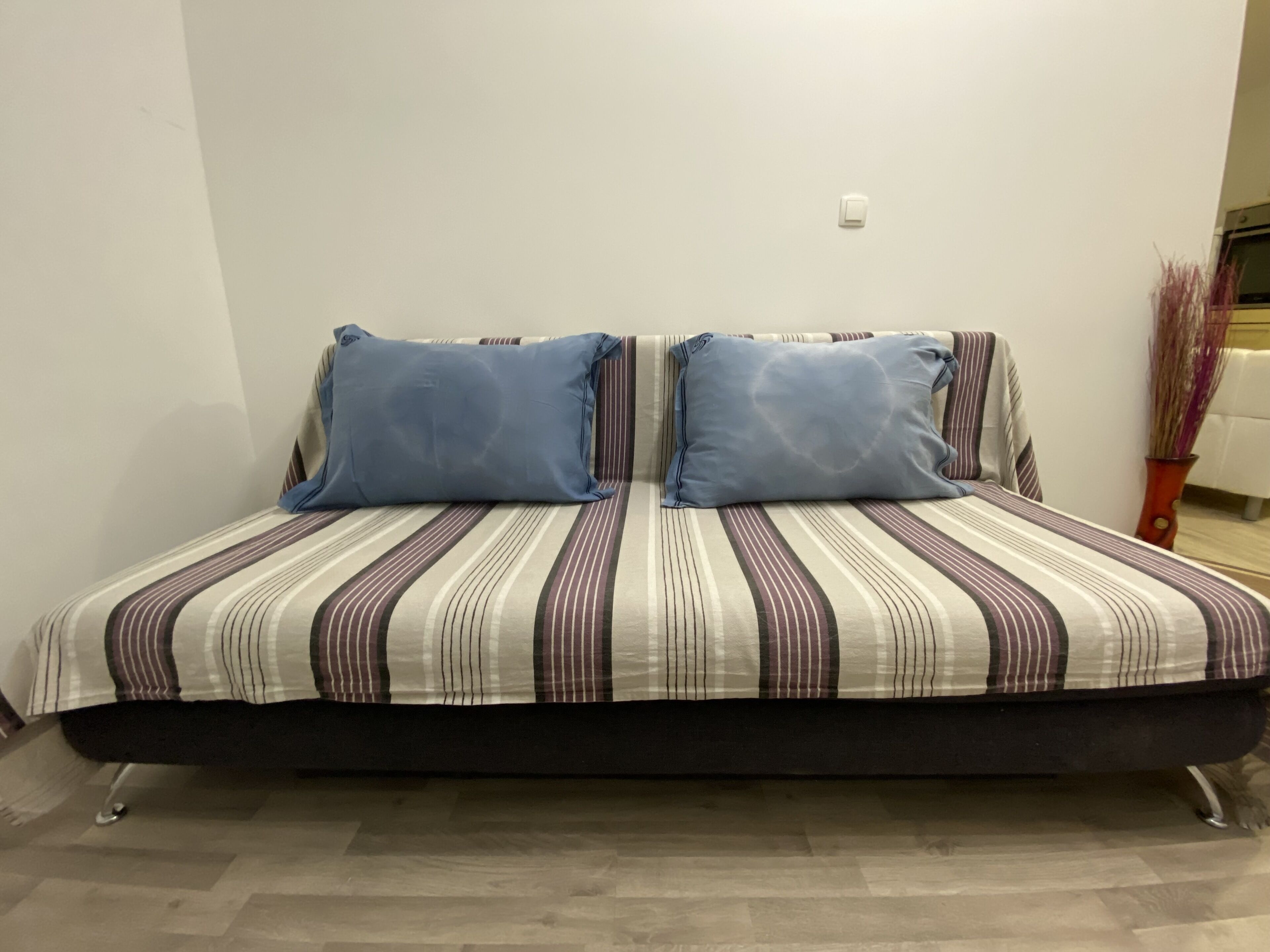 A bed with striped bedding and two pillows, a wall switch, and a vase with dried plants.
