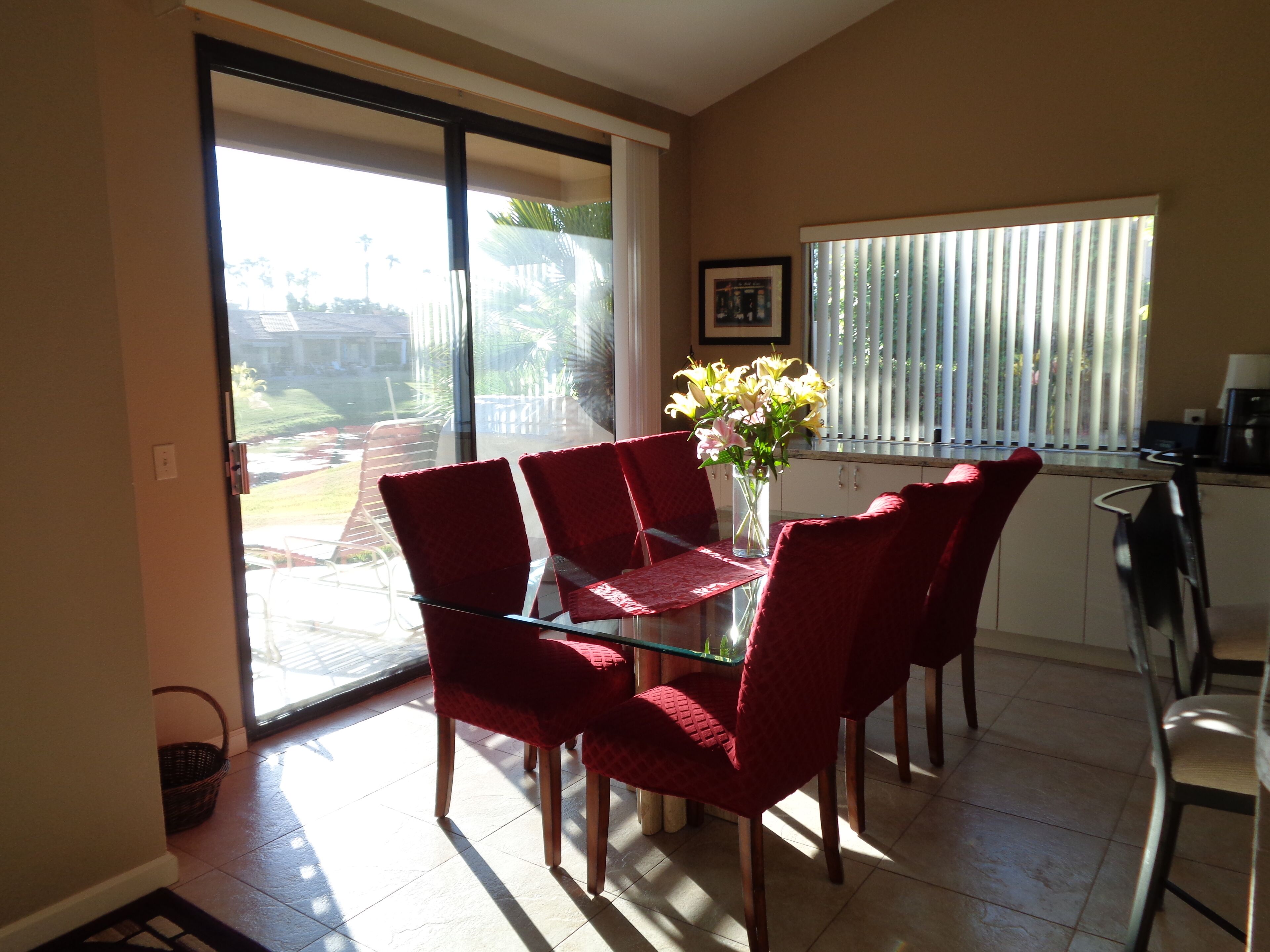 A dining area with a glass table and red chairs, a vase with flowers, and a view through a sliding glass door to a garden.