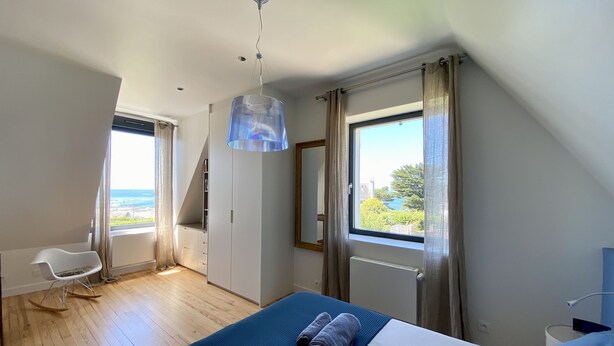 A bedroom with a bed, a rocking chair, a wardrobe, a window with a view, and a hanging light fixture. - Grande Villa, vue Mer, à Bénodet (Bénodet)