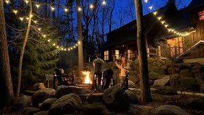 A rustic cabin with a fire pit surrounded by rocks and trees, string lights, and a wooden deck.