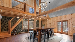 A spacious wooden interior with a high ceiling, a central dining table with stools, and large windows allowing natural light.