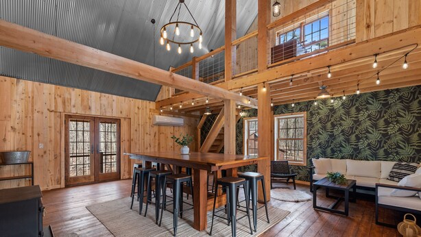 A spacious wooden interior with a high ceiling, a central wooden table with stools, a sofa, and a staircase.