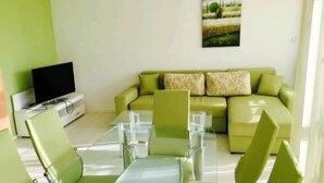 A modern living room with a green sofa, a flat-screen TV, and a dining area with a glass table and chairs. - Stylish 1-bed Apartment in Sunny Beach (Sunny Beach)