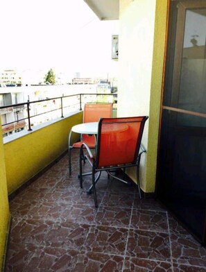 A small balcony with two orange chairs and a table, a tiled floor, and a view of buildings in the background.