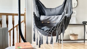 Puupalkkiin ripustettu riipputuoli, puinen tuoli, yöpöytä, jossa on kirja, punottu kori ja seinälle kiinnitetty kyltti.
