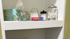 Ein Regal mit einer Packung Colgate Cavity Protection, einem Döschen Wattestäbchen und einer Schachtel Wattebäuschen. Darunter befinden sich gefaltete Handtücher in Dunkel- und Blautönen.