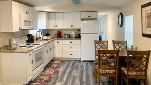 Une cuisine compacte avec des meubles blancs, un four à micro-ondes, une cuisinière et un réfrigérateur. On y trouve également un coin repas avec des chaises et une table en bois.