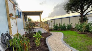 Un jardin avec un chemin de gravier, une terrasse en bois et un bâtiment dont la façade est recouverte de tôles ondulées.