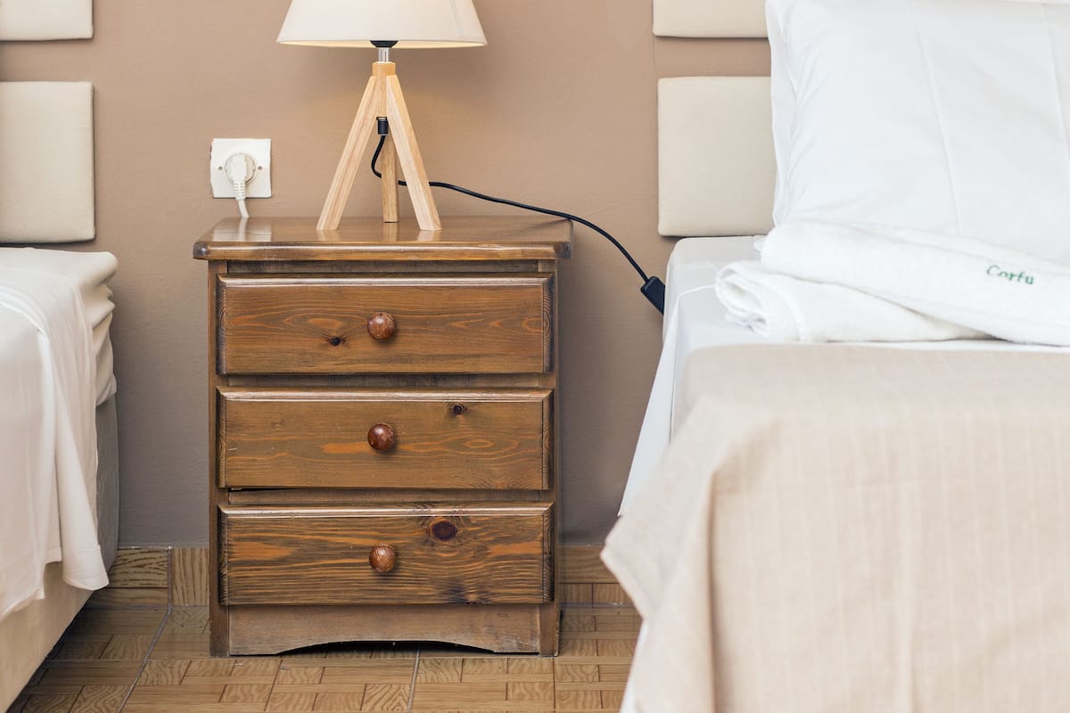 Ein ordentlich bezogenes Bett mit weißer Decke und Kissen, ein Nachttisch aus Holz mit einer Lampe und eine Steckdose.