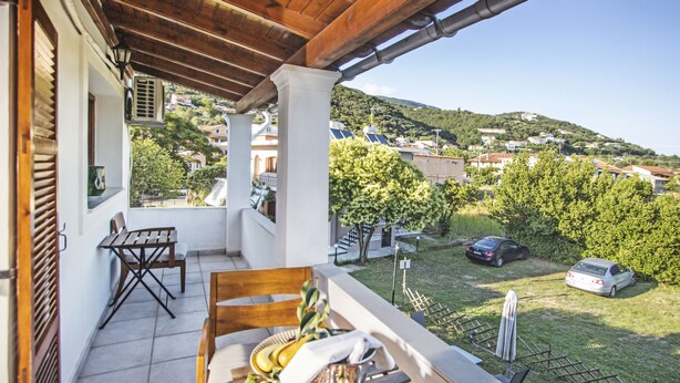 A balcony with a table set for dining, a chair, and a view of a hillside with houses and greenery. - NIKOLOUZOS SUNNY ESTATE (Corfu)