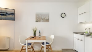Una habitación pequeña y moderna con una mesa de madera y dos sillas, una planta en maceta, un reloj en la pared y una zona de cocina con armarios blancos.