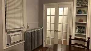 Une pièce avec un radiateur, une fenêtre équipée de stores, une porte avec des panneaux de verre et une table en bois avec des chaises.