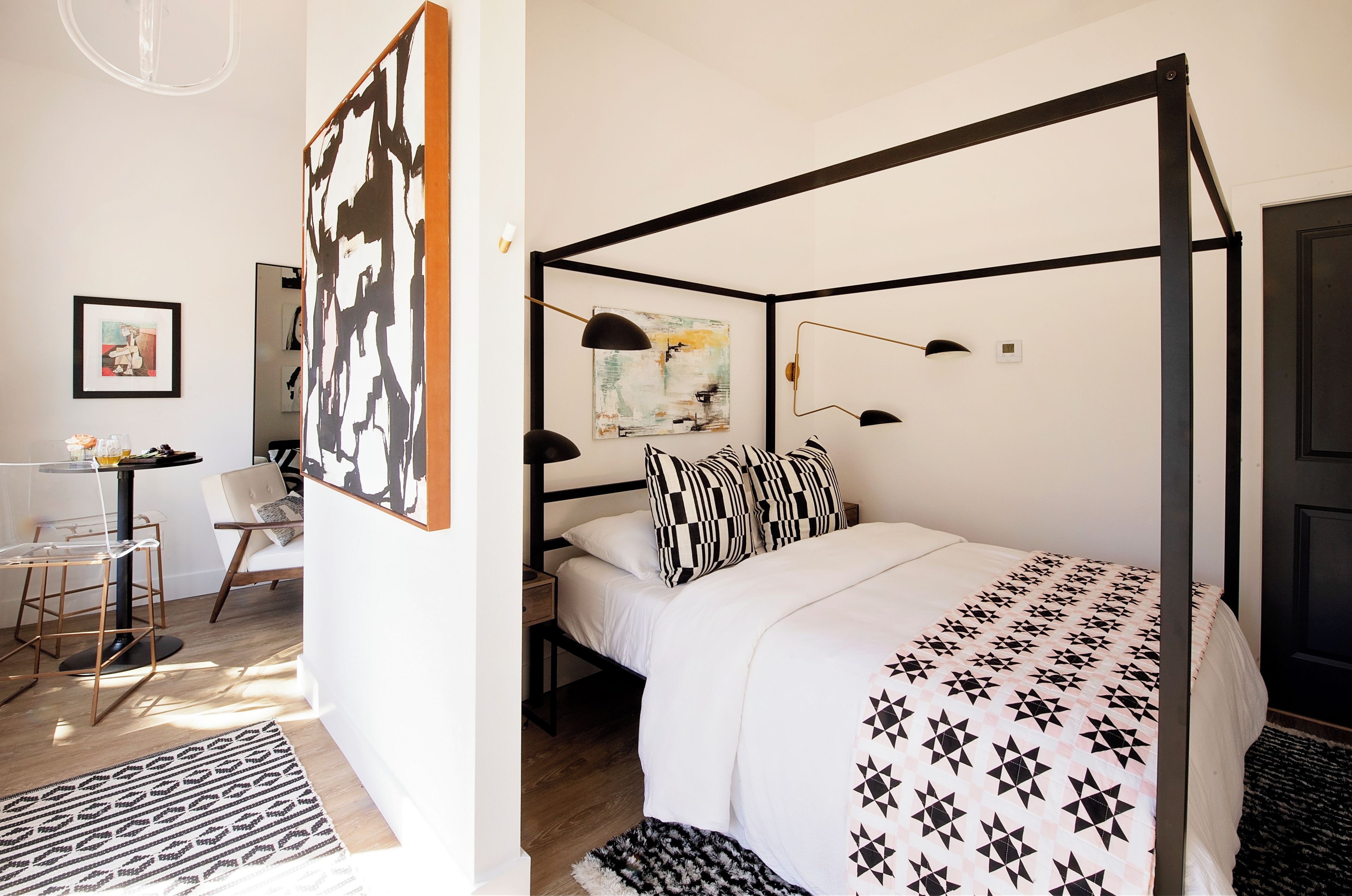 Una cama con dosel con una colcha de estampado geométrico, un cuadro abstracto blanco y negro en la pared, y un pequeño escritorio con una silla.