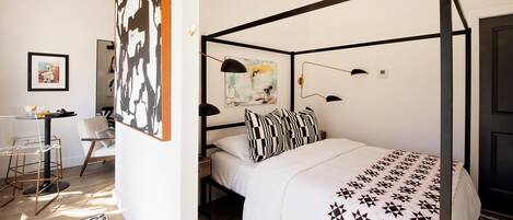 Una cama con dosel con una colcha de estampado geométrico, un cuadro abstracto blanco y negro en la pared, y un pequeño escritorio con una silla.