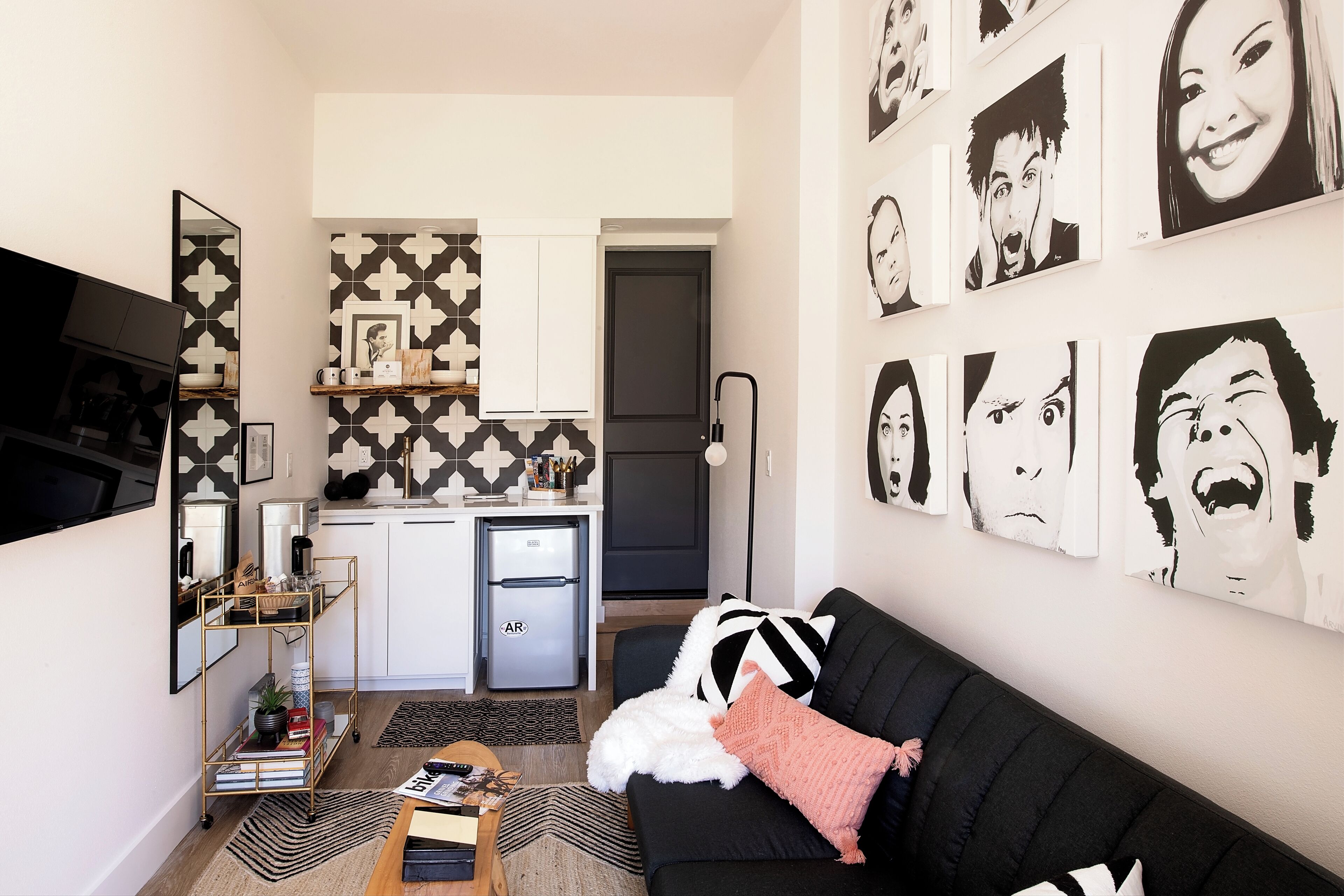 A compact living space with a kitchenette, a black sofa with a white and black pillow, and a wall adorned with black and white artwork.