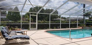 A covered outdoor swimming pool area with lounge chairs and a table, surrounded by a glass enclosure.
