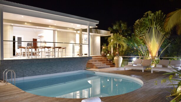 Een modern buitenzwembad met een houten terras, een lounge met witte stoelen, en uitzicht op de binnenruimte.