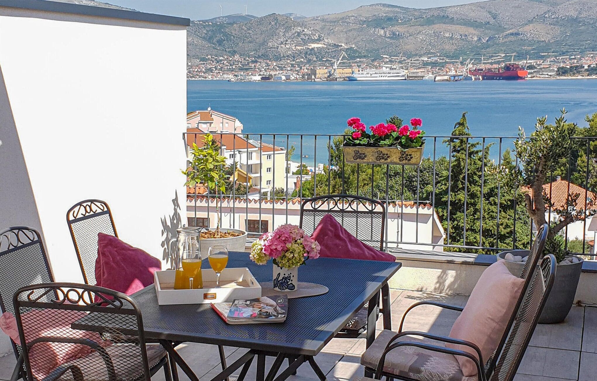 A balcony with a table set for two, overlooking a coastal town and the sea.
