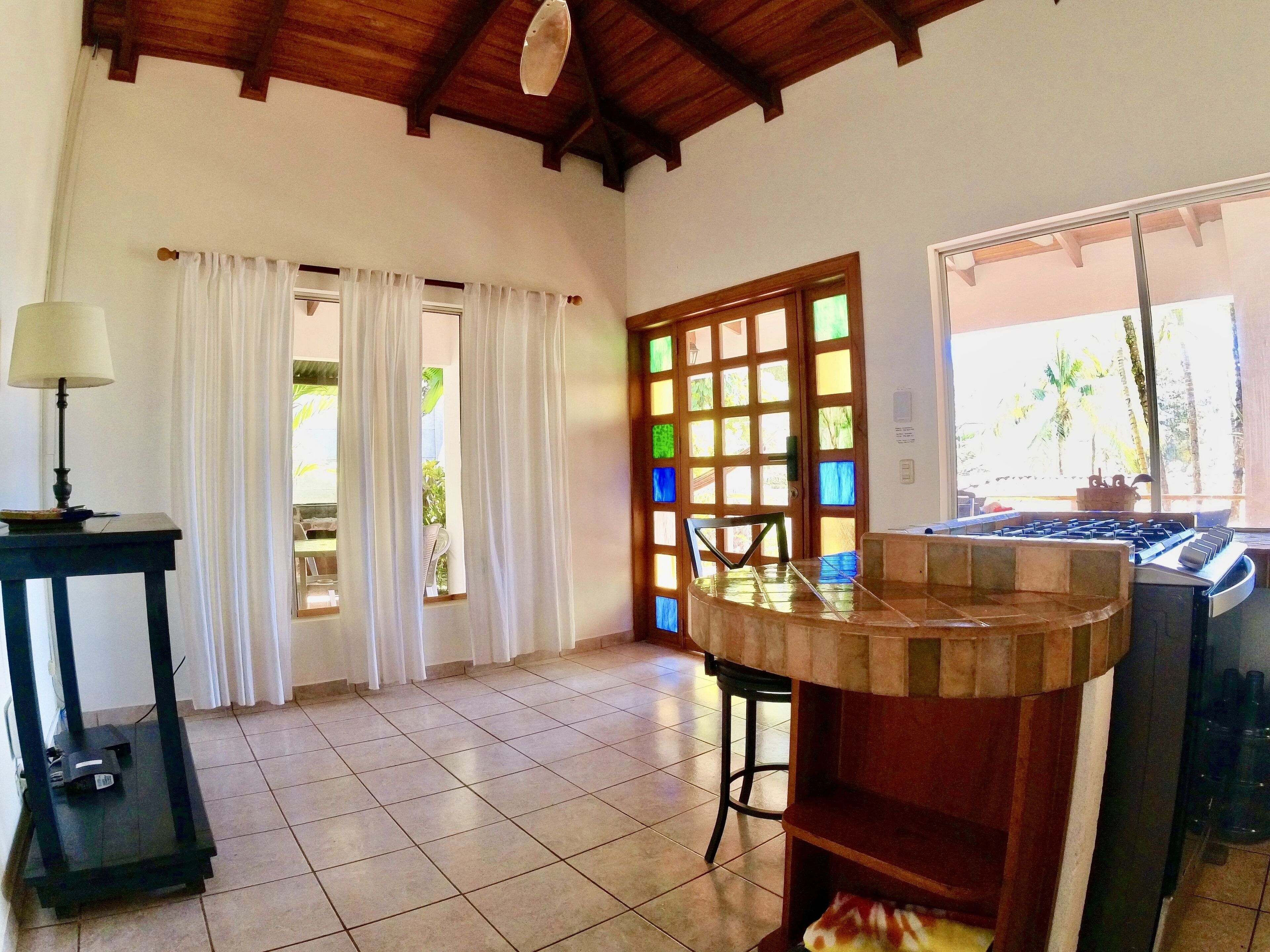 Una cocina con estufa a gas, una mesa y una silla. Se observa una ventana con cortinas y una puerta con vidrieras.