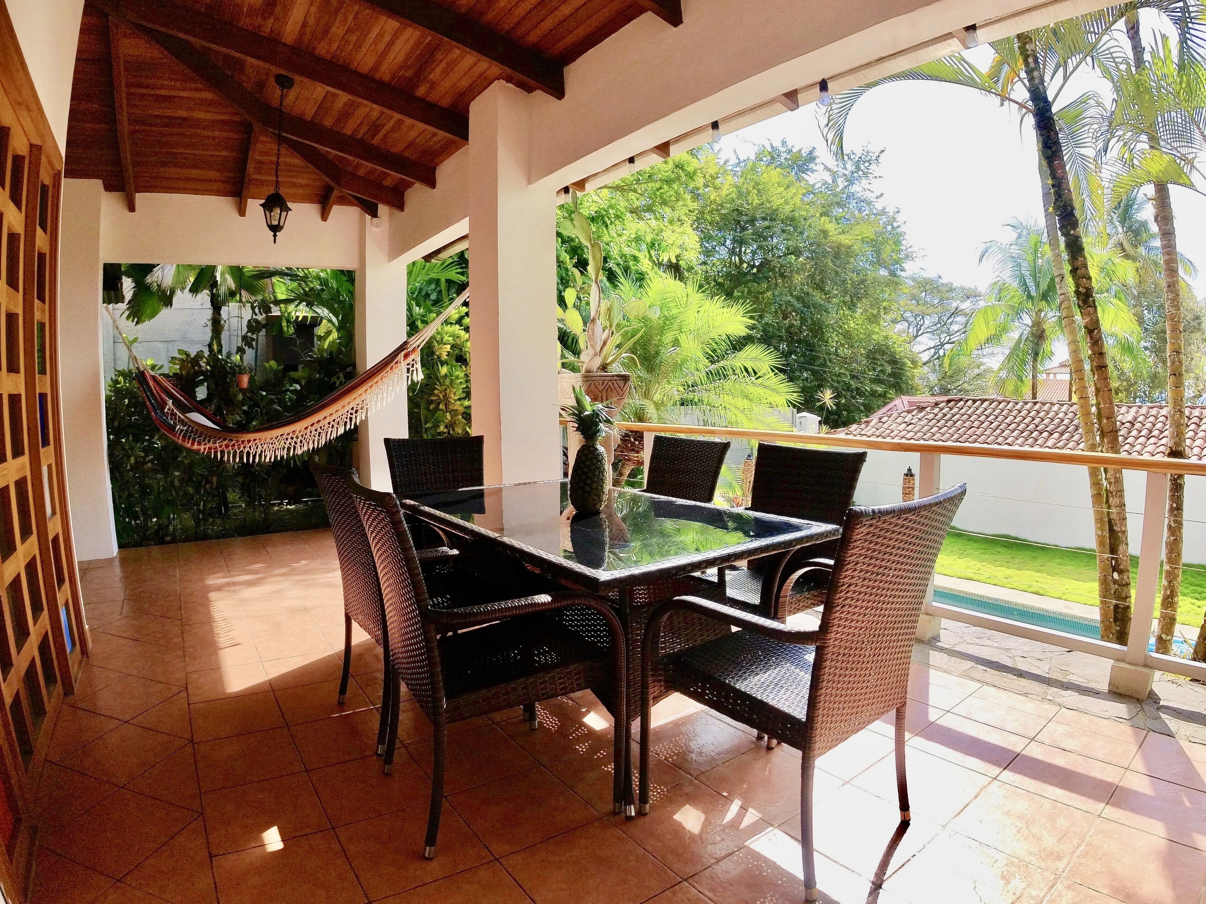 A covered patio with a glass table and wicker chairs, surrounded by lush greenery and a view of a swimming pool.