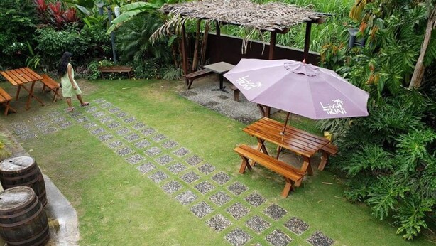 A garden area with a wooden bench, a table, and a purple umbrella. There are stone tiles on the ground and a thatched structure in the background. - Green Space, Santa Cruz Valley, Trinidad 2 (La Pastora Settlement)