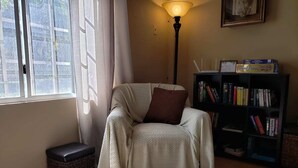 A cozy reading nook with a cushioned chair, a standing lamp, a bookshelf, and a window with curtains. - Green Space, Santa Cruz Valley, Trinidad 2 (La Pastora Settlement)