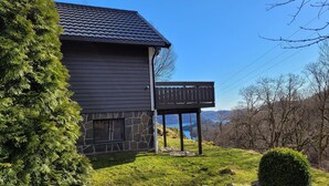Uma casa de dois andares com alicerce de pedra e revestimento escuro, uma varanda com guarda-corpo de madeira e vista para um lago e montanhas ao fundo.