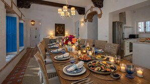 A dining area with a long table set for a meal, featuring a chandelier, wicker chairs, and a view of the kitchen. - Amazing 6 bed villa with private pool and views (Almuñecar)