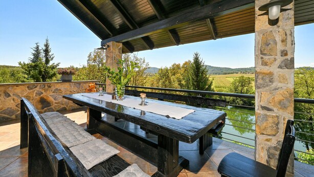 Une terrasse extérieure avec une table et des bancs en pierre, entourée de murs de pierre et offrant une vue sur un lac et des collines.