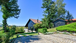 Une maison de campagne en bois, avec un toit recouvert de tuiles rouges, entourée d’une végétation luxuriante et bordée d’une allée pavée.