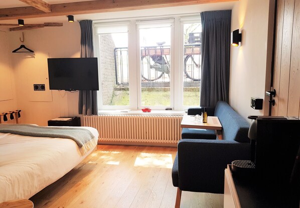A modern hotel room with a bed, a desk, a chair, a TV, and a window with a view of bicycles outside. - Stay in our centrally located 18th century house. Contactless stay possible. (Amsterdam)