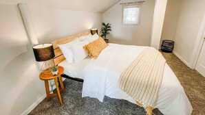 Ein Schlafzimmer mit einem großen Bett, einem Nachttisch mit Lampe, einer kleinen Topfpflanze und einem Fenster mit Vorhängen.