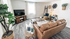 Ein modernes Wohnzimmer mit einer Ledersofa, einem Fernseher auf einem Holzbodenständer, einem Deckenventilator und einer großen Pflanze.