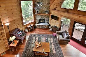 Interior acogedor de una cabaña con chimenea de piedra, muebles de madera y ventanas amplias que dan al exterior.