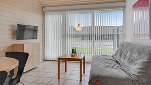 Uma sala de estar moderna com uma televisão de tela plana, um sofá cinza, uma mesa de jantar de madeira com cadeiras e uma porta de vidro deslizante que dá para uma área externa.